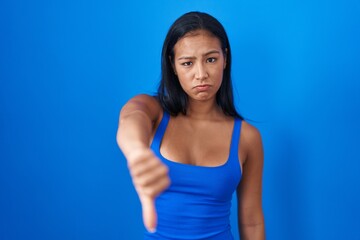 Fototapeta na wymiar Hispanic woman standing over blue background looking unhappy and angry showing rejection and negative with thumbs down gesture. bad expression.