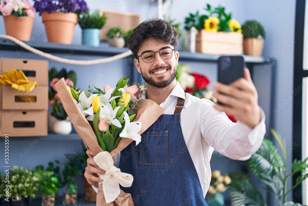 Wall mural young hispanic man florist make selfie by smartphone holding flowers at florist shop