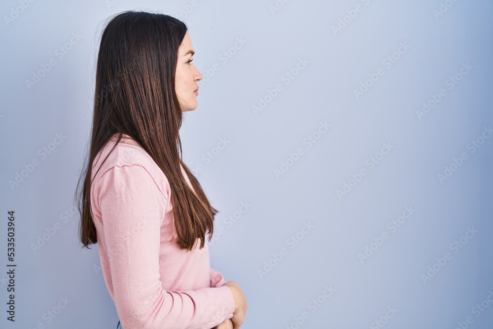 Poster young brunette woman standing over blue background looking to side, relax profile pose with natural 