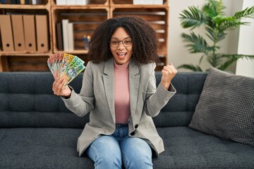 Young african american woman holding australian dollars at consultation office screaming proud,...