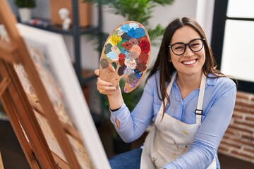 Young brunette woman holding painter palette at art studio looking positive and happy standing and smiling with a confident smile showing teeth