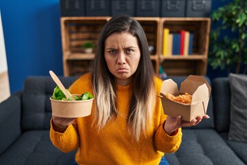 Young hispanic woman holding healthy salad and fried chicken wings depressed and worry for...