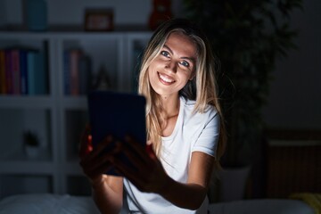 Young blonde woman using touchpad sitting on bed at bedroom