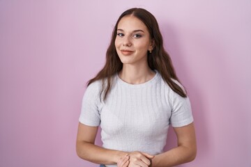 Fototapeta premium Young hispanic girl standing over pink background with hands together and crossed fingers smiling relaxed and cheerful. success and optimistic