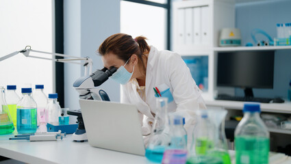 Middle age hispanic woman wearing scientist uniform and medical mask using microscope at laboratory