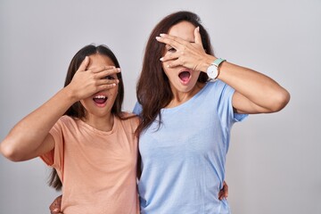 Young mother and daughter standing over white background peeking in shock covering face and eyes with hand, looking through fingers with embarrassed expression.