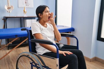Young hispanic woman sitting on wheelchair at physiotherapy clinic clueless and confused with open...