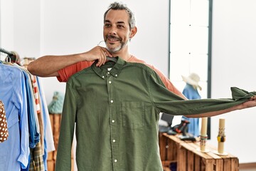 Middle age grey-haired man customer smiling confident holding shirt of rack at clothing store