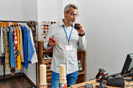 Middle age grey-haired man shop assistant using smartphone drinking mate at clothing store