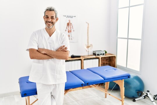 Middle age hispanic therapist man working at pain recovery clinic happy face smiling with crossed arms looking at the camera. positive person.