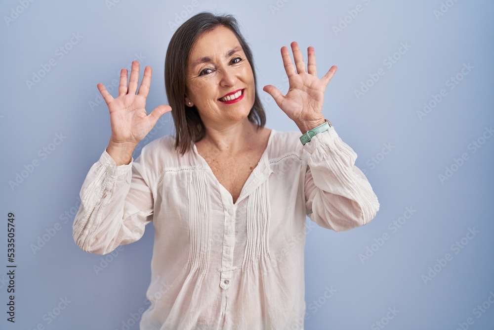 Sticker Middle age hispanic woman standing over blue background showing and pointing up with fingers number ten while smiling confident and happy.