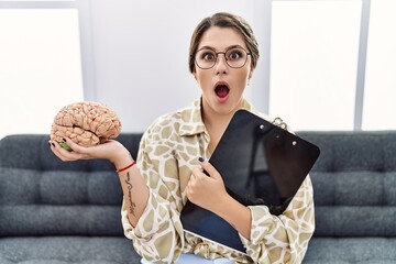 Young hispanic woman holding brain working at psychology clinic afraid and shocked with surprise...