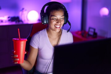 Young beautiful hispanic woman streamer playing video game drinking beverage at gaming room