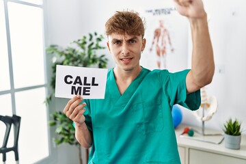 Young caucasian physio man holding call me banner at the clinic annoyed and frustrated shouting...