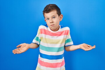 Young caucasian kid standing over blue background clueless and confused expression with arms and hands raised. doubt concept.