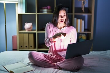 Young hispanic woman using computer laptop on the bed pointing with finger surprised ahead, open mouth amazed expression, something on the front