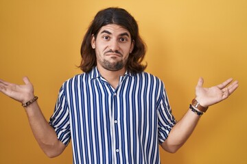 Hispanic man with long hair standing over yellow background clueless and confused expression with...