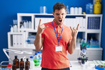 Young hispanic man working at scientist laboratory shouting with crazy expression doing rock symbol...