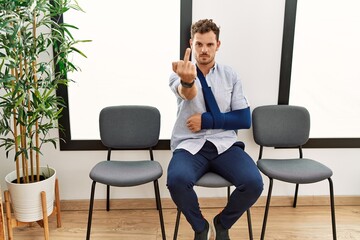 Handsome young man sitting at doctor waiting room with arm injury showing middle finger, impolite and rude fuck off expression