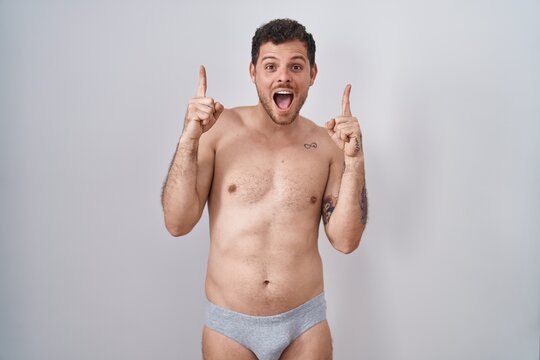 Young hispanic man standing shirtless wearing underware smiling amazed and surprised and pointing up with fingers and raised arms.