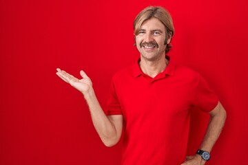 Caucasian man with mustache standing over red background smiling cheerful presenting and pointing with palm of hand looking at the camera.