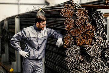 A metallurgy worker stands next to iron and steel bars in the facility and looks at them.