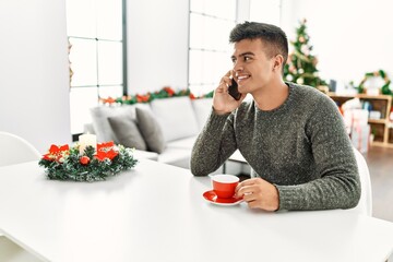 Young hispanic man drinking coffee and talking on the smartphone sitting by christmas tree at home