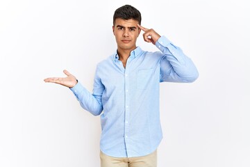 Young hispanic man wearing business shirt standing over isolated background confused and annoyed with open palm showing copy space and pointing finger to forehead. think about it.
