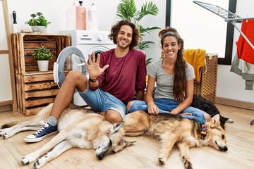 Young hispanic couple doing laundry with dogs showing and pointing up with fingers number five while smiling confident and happy.