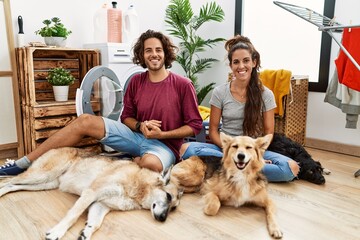 Young hispanic couple doing laundry with dogs with hands together and crossed fingers smiling relaxed and cheerful. success and optimistic