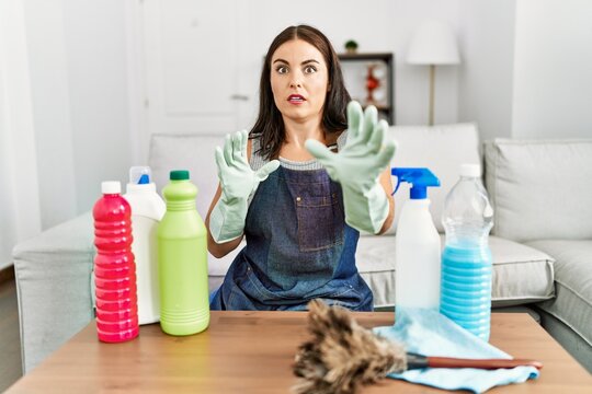Young brunette woman wearing cleaner apron and gloves cleaning at home afraid and terrified with fear expression stop gesture with hands, shouting in shock. panic concept.