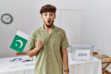 Young arab man at political campaign election holding pakistan flag scared and amazed with open...