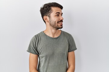 Young hispanic man with beard wearing casual t shirt over white background looking away to side with smile on face, natural expression. laughing confident.