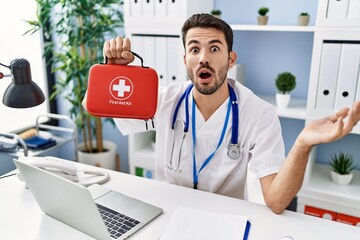 Young hispanic doctor man holding first aid kit scared and amazed with open mouth for surprise,...