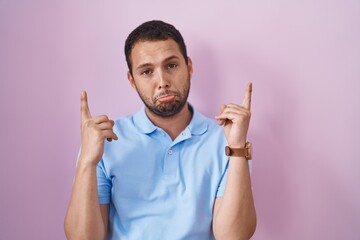 Hispanic man standing over pink background pointing up looking sad and upset, indicating direction with fingers, unhappy and depressed.