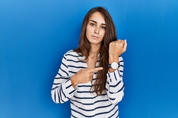 Young hispanic woman standing over blue isolated background in hurry pointing to watch time, impatience, looking at the camera with relaxed expression