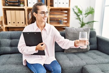 Young hispanic woman holding water having psychologist session at clinic