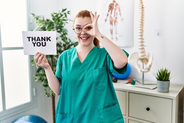 Young redhead physiotherapist woman working at pain recovery clinic holding thank you banner smiling happy doing ok sign with hand on eye looking through fingers