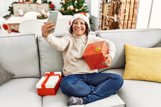 Middle age hispanic woman make selfie by the smartphone sitting on the sofa by christmas tree at home.