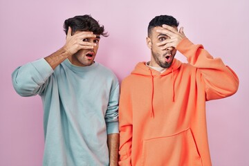 Young hispanic gay couple standing over pink background peeking in shock covering face and eyes...