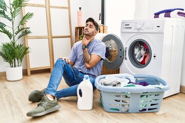 Young hispanic man putting dirty laundry into washing machine touching painful neck, sore throat for flu, clod and infection