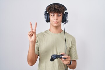 Hispanic teenager playing video game holding controller smiling with happy face winking at the camera doing victory sign with fingers. number two.