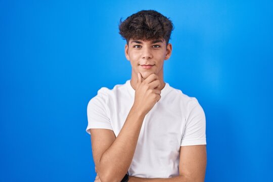 Hispanic teenager standing over blue background looking confident at the camera smiling with crossed arms and hand raised on chin. thinking positive.