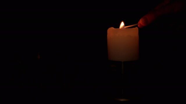 Hand Lights a Candle Wick in a Candlestick using a Match on a Black Background. Flame burning candle on the background of dry withered flowers, roses. Concept of memory, ritual, sorrow, anniversary.