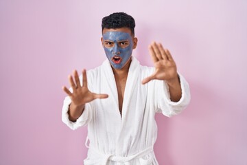 Young hispanic man wearing beauty face mask and bath robe doing stop gesture with hands palms, angry and frustration expression