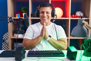 Young hispanic man playing video games praying with hands together asking for forgiveness smiling...