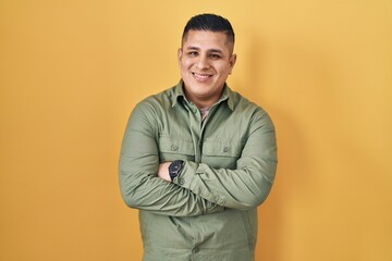 Hispanic young man standing over yellow background happy face smiling with crossed arms looking at the camera. positive person.