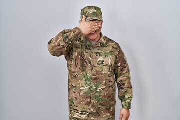 Hispanic young man wearing camouflage army uniform smiling and laughing with hand on face covering eyes for surprise. blind concept.