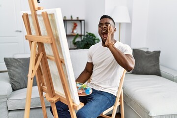 Young african man painting on canvas at home shouting and screaming loud to side with hand on mouth. communication concept.