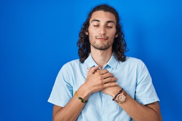 Young hispanic man standing over blue background smiling with hands on chest with closed eyes and...